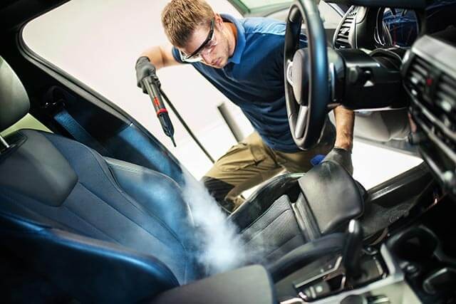 A contractor car valet using a steam cleaner on a car's interior, focusing on the driver's seat.