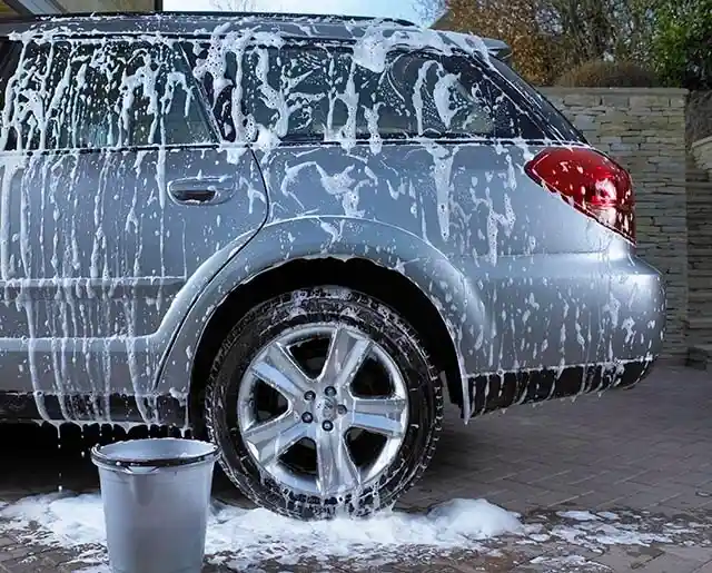 Silver car covered in soap suds, parked for a wash with a bucket nearby.