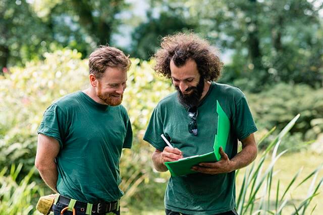 Two men in green shirts discussing in a garden, one holding a folder and pen.
