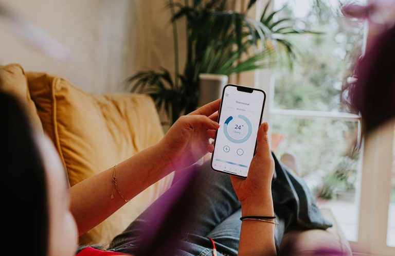 Person holding smartphone with thermostat app set to 24 degrees, on a couch.
