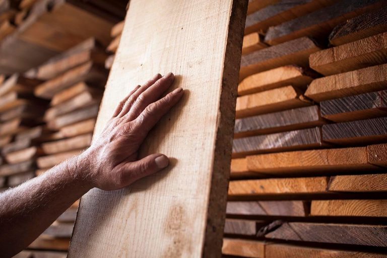 Close-up of a hand touching a smooth wooden plank, with stacks of wood in the background.