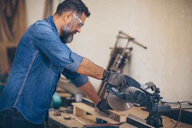 Wood worker using a miter saw to cut wood in a workshop, surrounded by tools.