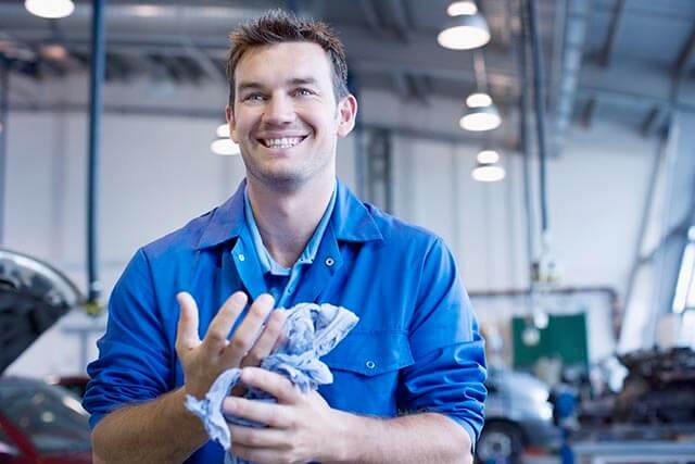 A mechanic in a blue uniform stands in a garage while smiling. He is holding a cleaning cloth. The background shows cars and garage equipment, with ceiling lights illuminating the space.