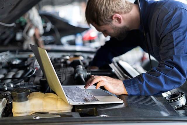 A mechanic in a blue jumpsuit is working under the hood of a car. He is using a silver laptop, which rests on the engine. His focus is on the engine, possibly diagnosing an issue or looking up repair information.
