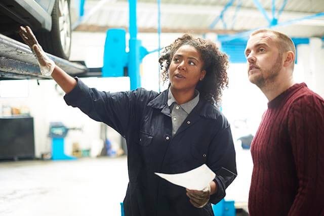 A mechanic in a workshop pointing towards a raised vehicle while explaining something to a man in a red sweater. The mechanic holds a piece of paper and wears a dark coverall. The workshop has blue equipment in the background.