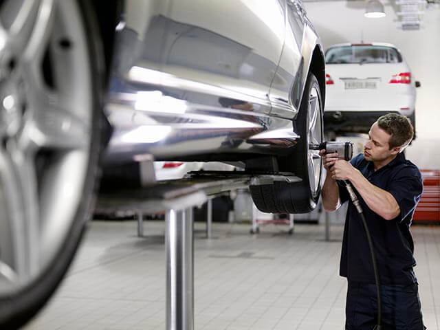 A mechanic works on a car elevated on a hydraulic lift, using a power tool to tighten or loosen bolts on a wheel. Another car is visible in the background, parked on an adjacent lift in a well-lit automotive repair shop.