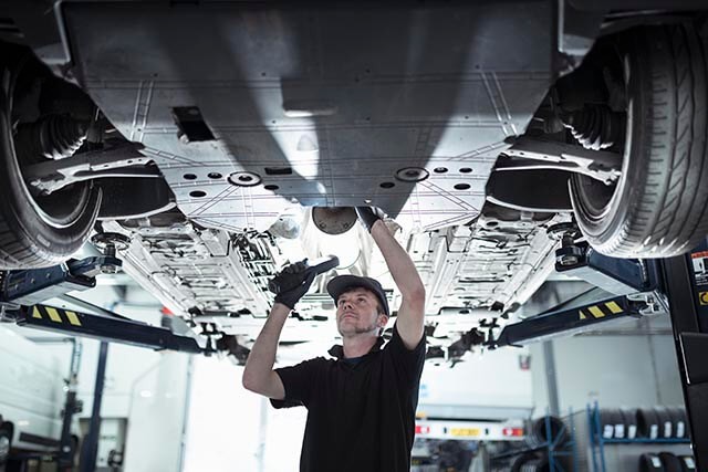A mechanic is inspecting the underside of a vehicle raised on a hydraulic lift in a garage. He is wearing a cap, gloves, and a dark work shirt. The workshop environment is well-lit, with various tools and equipment in the background.