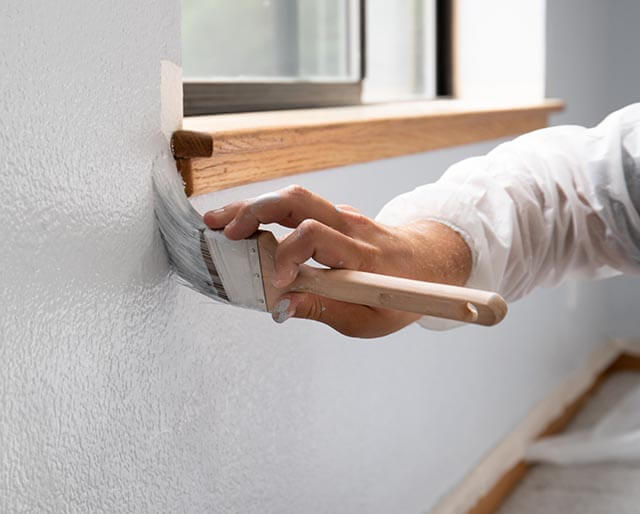 A person wearing a white sleeve is painting a light-colored wall with a paintbrush. The window in the background has a wooden frame. The room appears to be well-lit.