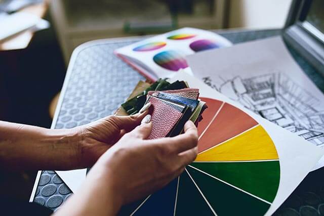 A person holds a selection of fabric swatches in their hands, sitting on a dark table with a color wheel and an architectural sketch nearby. The scene suggests a design or planning process.