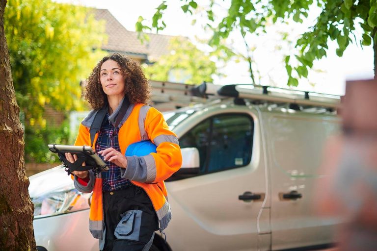 Person holding tablet and hard hat walking outdoors near a white utility van.