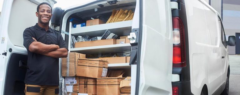 A person stands beside an open commercial van filled with various packages and envelopes, showcasing the benefits of reliable van insurance. The white van features shelves neatly arranged with parcels. Smiling in a black shirt and pants, the person confidently crosses their arms.