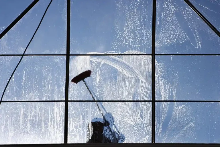 Window cleaner cleaning a large window with a brush, soap suds and clear blue sky visible.