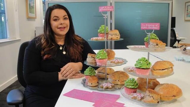 A woman seated at a table with multiple trays of baked goods, including scones and green frosted cupcakes. She is smiling, and there are small pink signs and cards on the table next to her. The setting appears to be an office or meeting room.