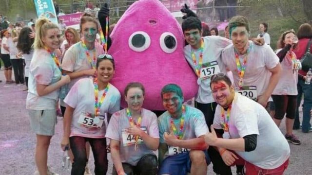 A group of people in colorful powder-stained outfits pose with a person in a large pink character costume. They are outdoors at a fun run event, wearing race numbers and rainbow lanyards. The ground is dusted with colored powder.