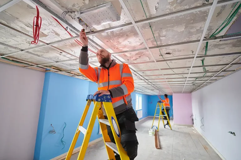 Workers installing a suspended ceiling in a room with ladders and tools, large windows letting in light.