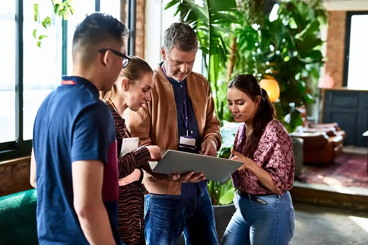 Four people stand indoors, focused on a laptop held by a man wearing a brown jacket. They are in a well-lit room with large windows and lush green plants. The group appears to be engaged in a discussion.