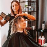 Hairstylist drying a woman’s hair in a salon. The client is wearing a black cape.