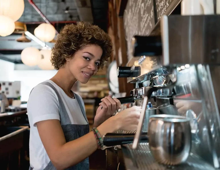A woman prepares coffee behind the counter in a coffee shop.