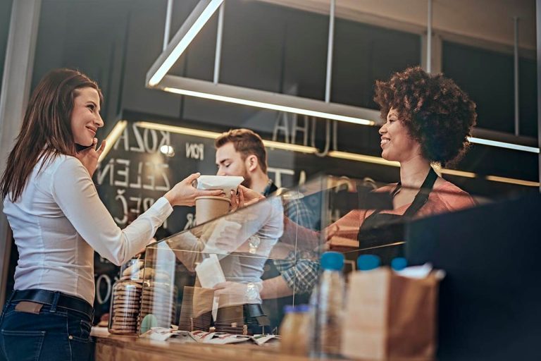 Woman at café counter receiving a wrapped dessert or pastry from a smiling barista, a man preparing a drink in the background.