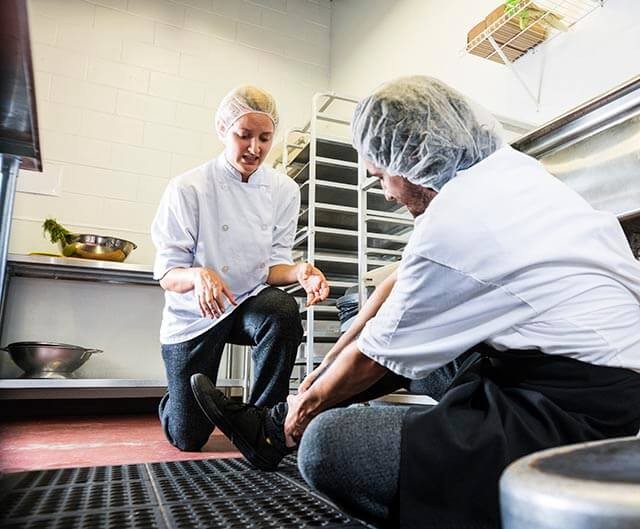Kitchen worker knelt on the floor and holding their leg in pain, with another colleague offering assistance.