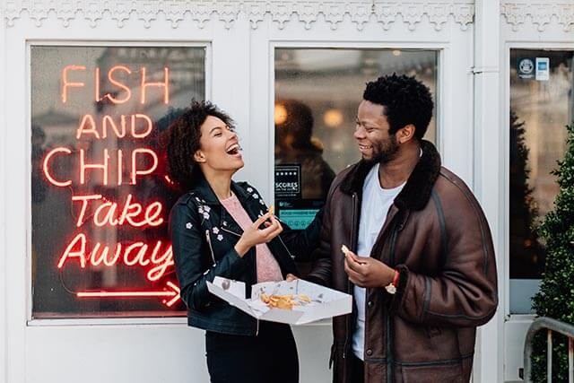 Two people outside a fish and chip shop, enjoying crispy chips with a neon sign in the background.