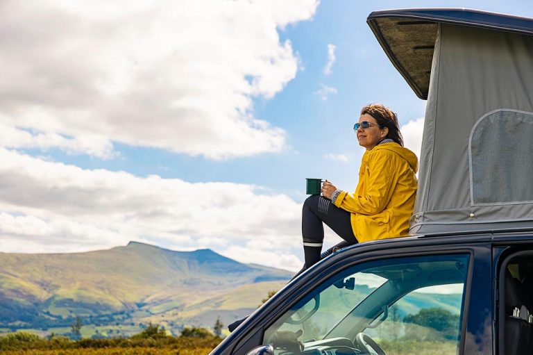 Person with a mug on a vehicle roof tent, enjoying a scenic landscape, with a mountain and a cloudy sky in the background.
