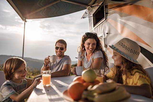A family of four sits at an outdoor table beside a camper. They are smiling and enjoying drinks. There are fruits like bananas and apples on the table. The setting is sunny, with hills in the background, and an awning provides shade.
