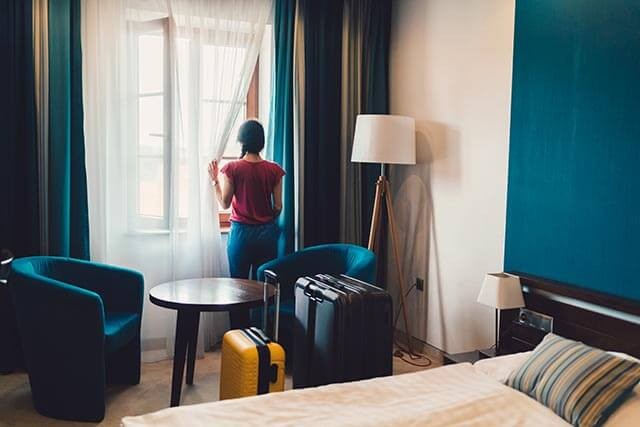 A hotel guest is standing by the window of a hotel room with suitcases beside the bed.
