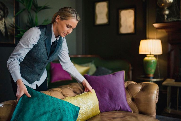 Hotel employee arranging pillows on a couch in a cozy, dimly lit hotel foyer.