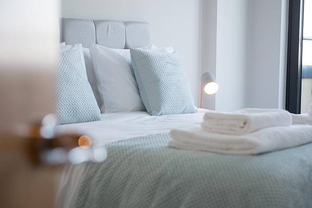 Neatly made bed in a cozy room with grey headboard, blue pillows, and white towels on the bed.