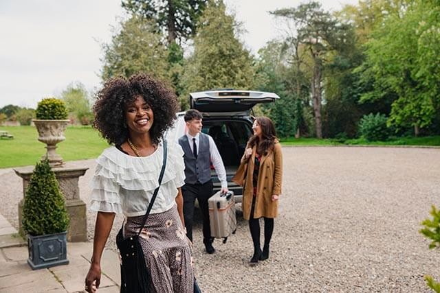 Three people standing in a hotel car park. A woman is smiling while an employee is helping with the suitcase.