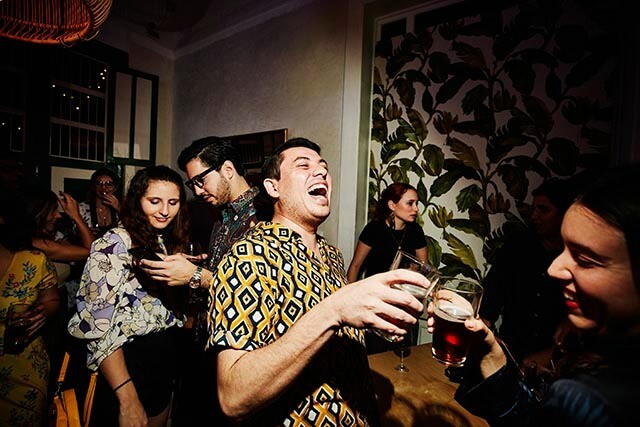 Man laughing while clinking his glass at a lively nightclub, surrounded by people chatting and dancing.