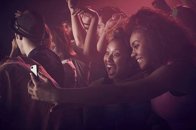 Two people smiling and taking a selfie in a lively nightclub with red lighting while people dance around them.