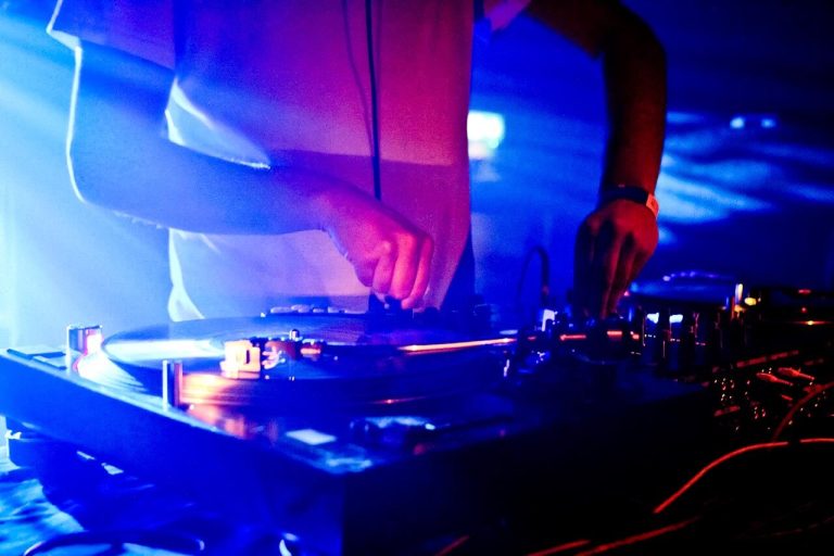 DJ adjusting controls on their console in a dimly lit nightclub with blue and pink lighting and smoke effects.