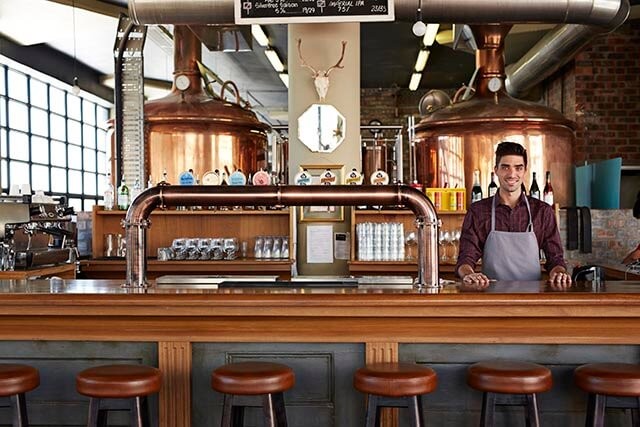 Bartender behind a bar with brewing tanks and beer taps in a brewery or pub setting with industrial-style decor.