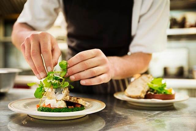 Chef wearing an apron garnishing a plated dish with fresh greens in a professional kitchen.