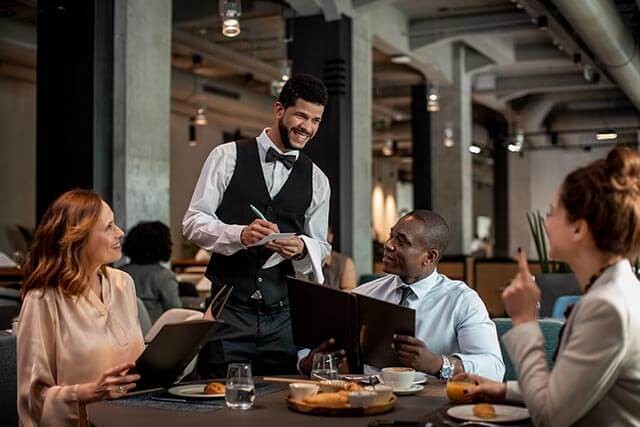 Waiter wearing a black vest and a bow tie taking orders from patrons in a modern restaurant while they hold menus.