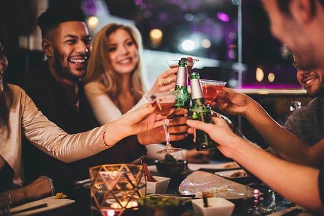Group of people raising drinks in a toast at a dimly lit table with food and a small candle.