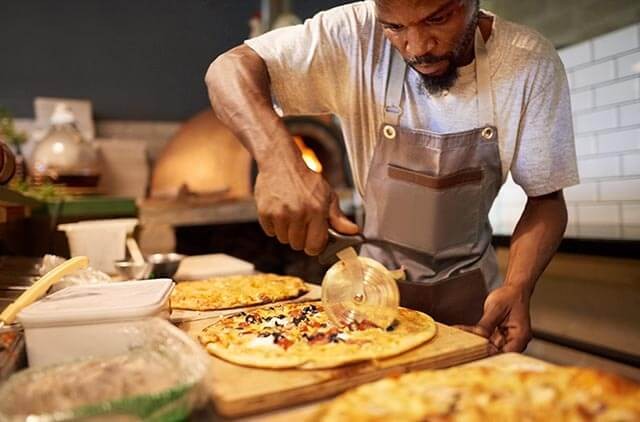 Takeway chef slicing freshly baked pizza in a kitchen with multiple pizzas on the counter.