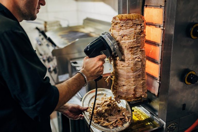 Employee shaving kebab meat from a rotating spit in a kitchen, serving it onto a plate for takeaway.