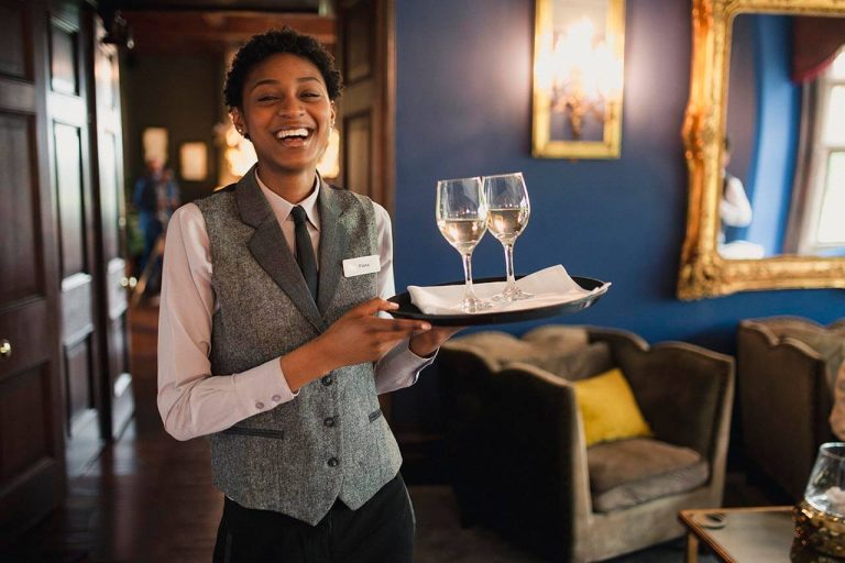 A smiling server in a vest and tie balancing a tray with wine glasses in a dimly lit, elegant room.