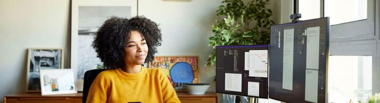 A woman seated at a desk, focused on her computer screen, surrounded by office supplies and a window in the background.