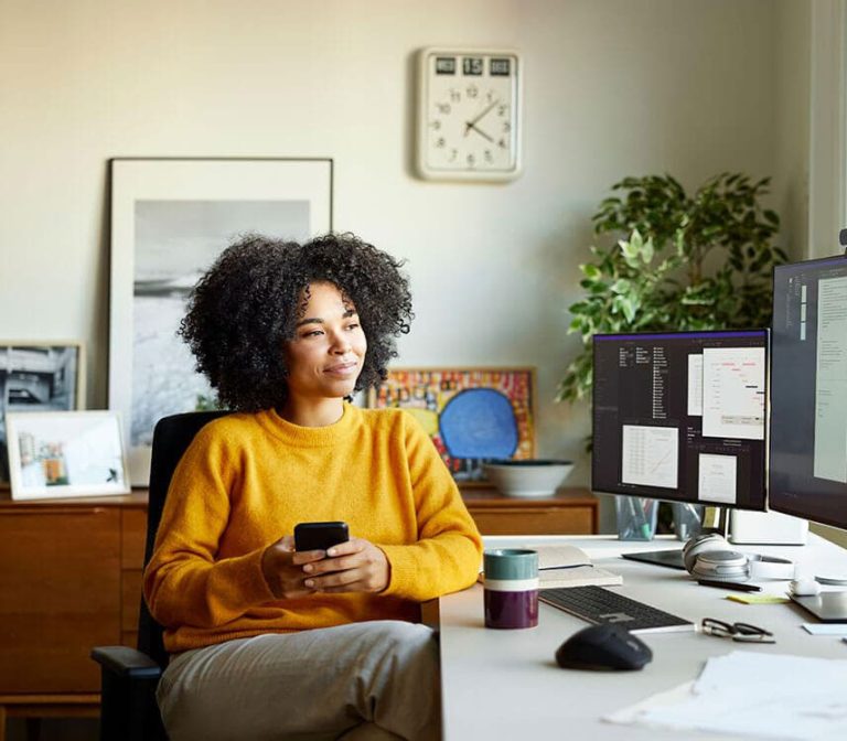 A woman seated at a desk, working on a computer while holding a cell phone in her hand.