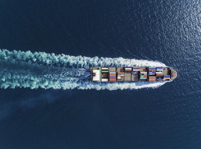 An aerial view of a cargo ship moving through deep blue water, leaving a white wake behind. The ships deck is loaded with multicolored shipping containers arranged in neat rows.