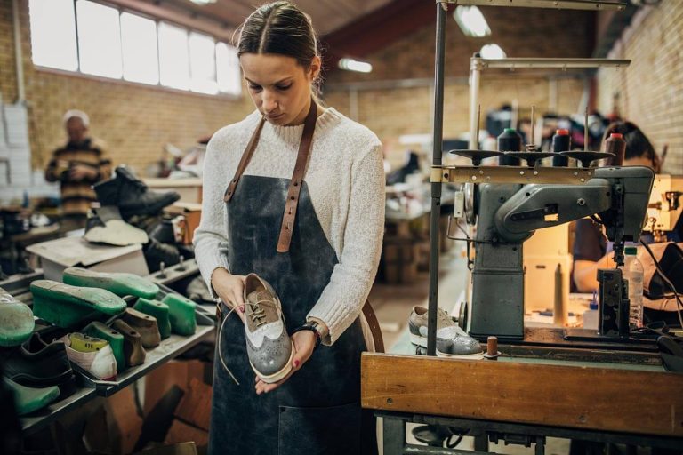Woman in an apron examining a shoe in a workshop, surrounded by materials and tools near a sewing machine.