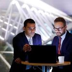 Two businessmen are looking at a laptop in a dimly lit, modern office setting.