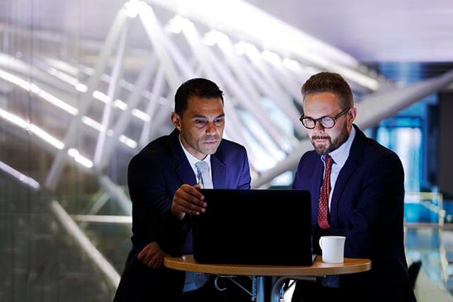 Two businessmen are looking at a laptop in a dimly lit, modern office setting.
