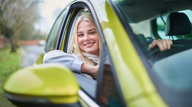 A smiling young person is taking a driving lesson. She is seated in the driver's seat with the window down.