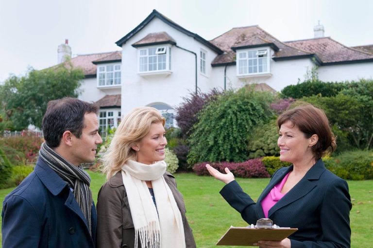 Female sales agent showing a house to a couple outdoors, holding a clipboard with a large white house in the background.