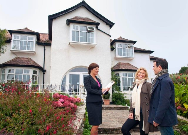Estate agent showing a house to two people outside, holding a clipboard, with flowers and a large house in the background.